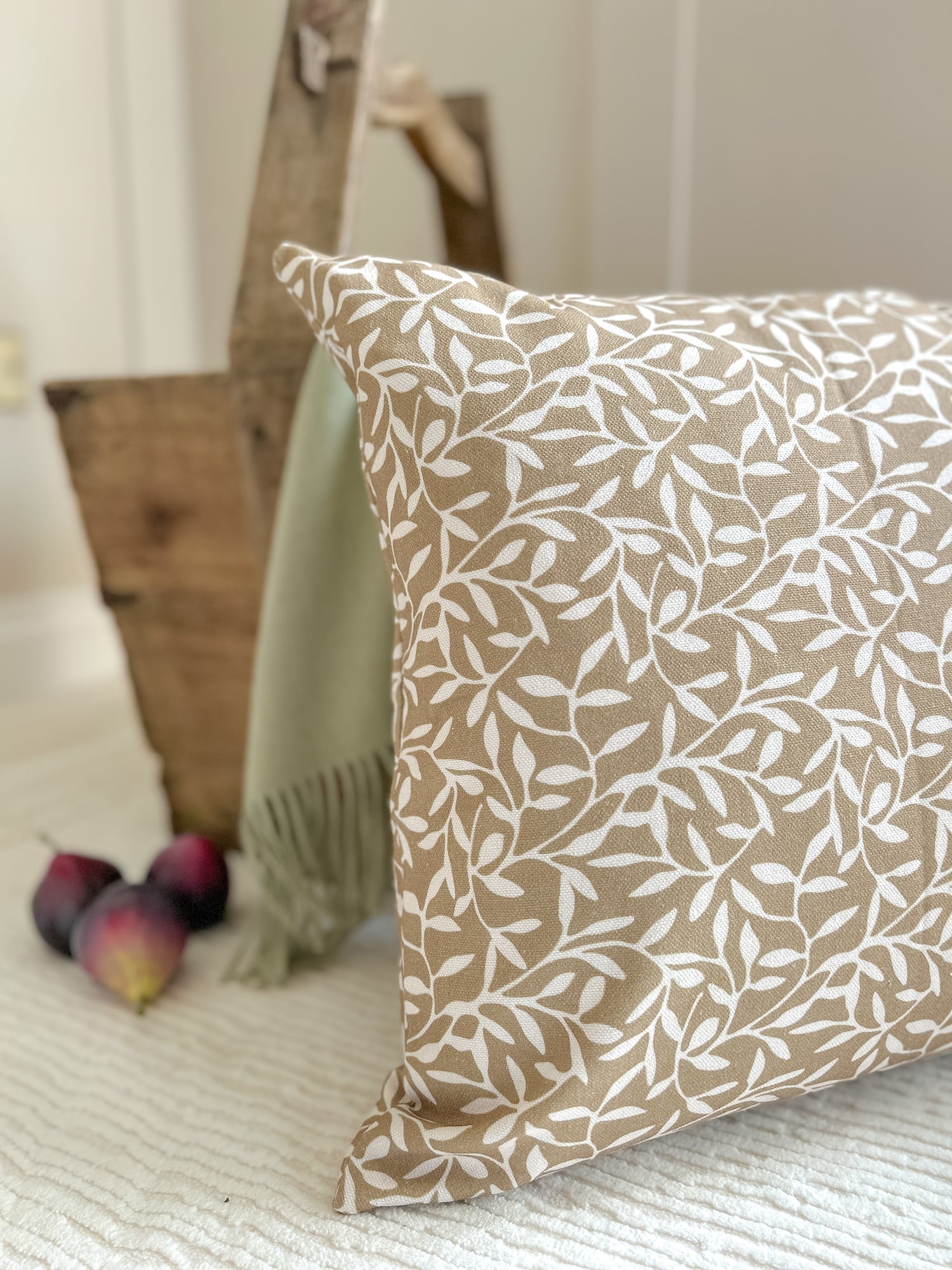 Olive green cushion with white leaf print, styled on a neutral textured background with figs and rustic accents for cozy home interiors.