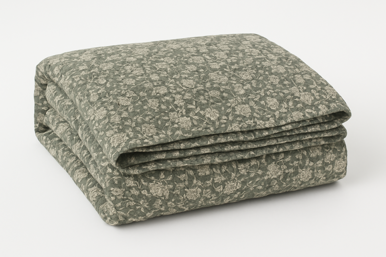 Sage green quilted throw folded neatly, floral patterned cotton blend blanket for layering on bed or sofa in calm, country-style interiors.