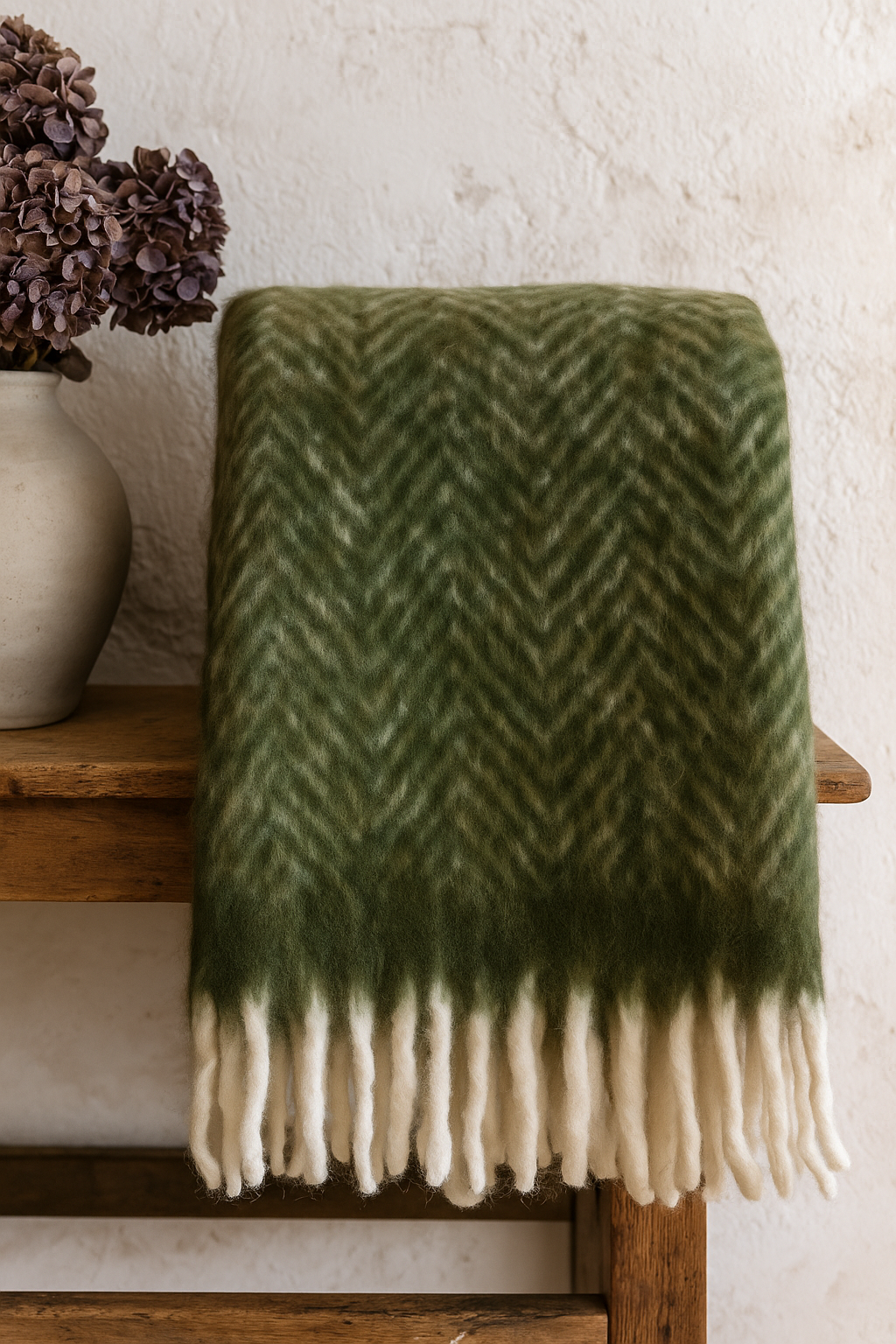 Green and white herringbone patterned blanket draped over a wooden stool with a vase of flowers in the background.
