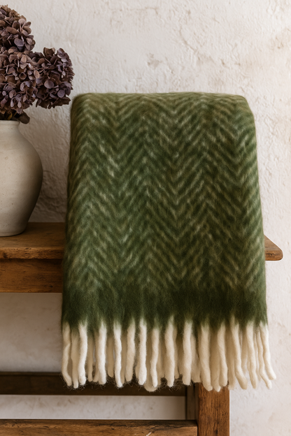 Green and white herringbone patterned blanket draped over a wooden stool with a vase of flowers in the background.