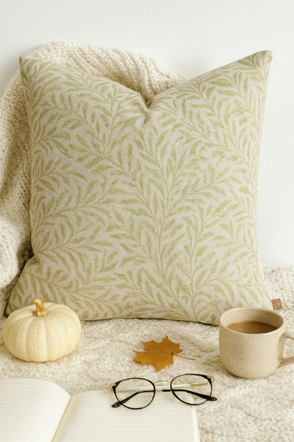 45x45cm square cushion cover in beige with olive green botanical leaf pattern, styled on a white background with cable knit throw, coffee mug, journal, glasses, and autumn accents.