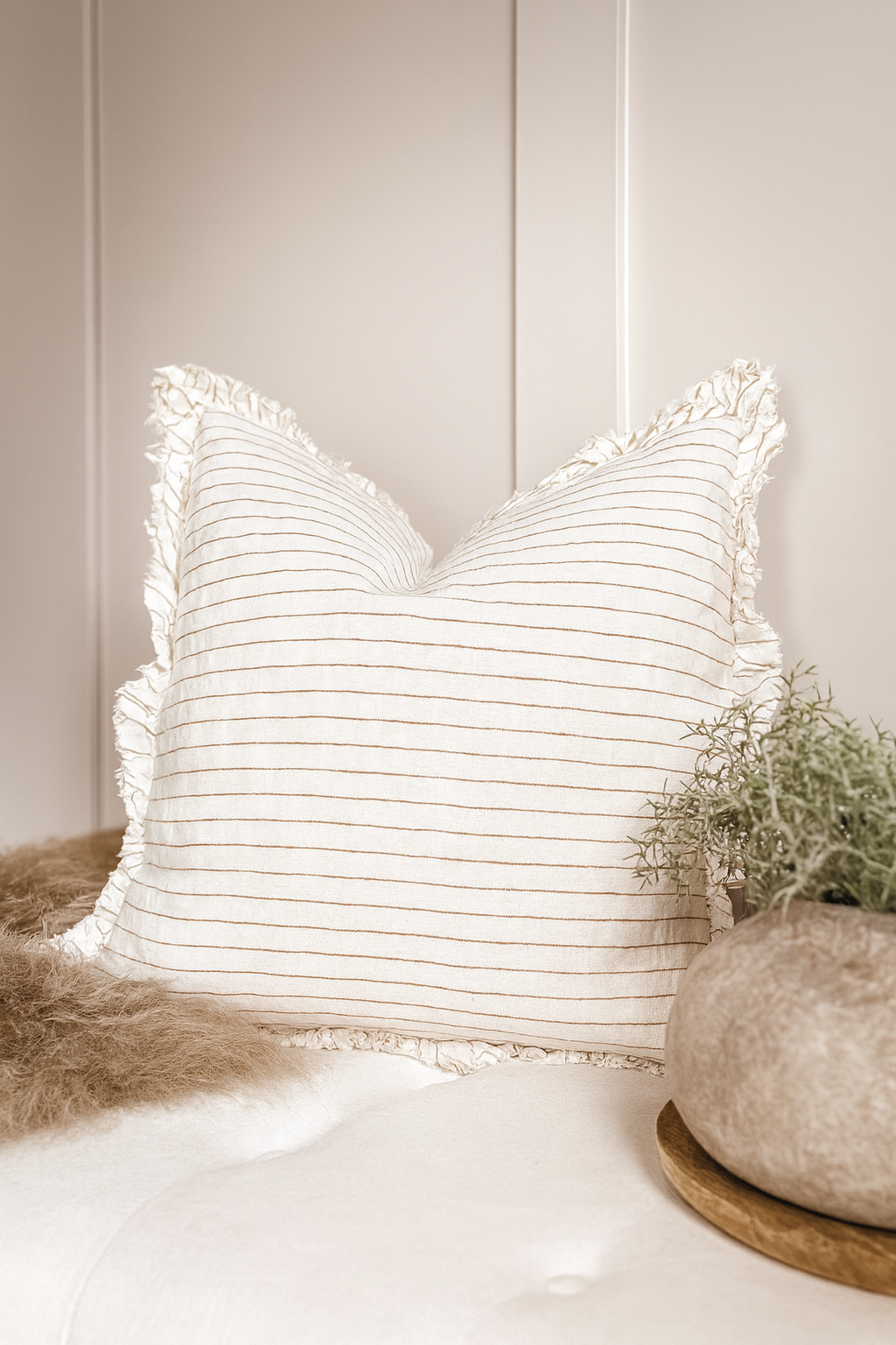 Neutral striped cushion with ruffled edges styled in a cosy country cottage setting, placed on a soft seat with textured throw and rustic décor