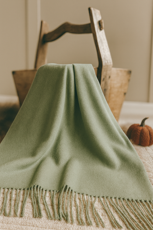 A soft sage green throw blanket with fringed edges draped over a rustic wooden basket, styled in a cozy neutral setting with a small autumnal pumpkin accent.